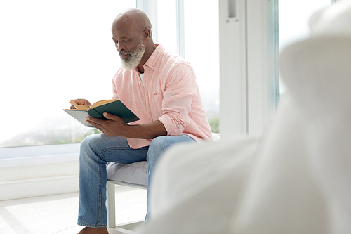 Side view of African American man reading a book and sitting on the bed in the bedroom. Authentic Senior Retired Life Concept