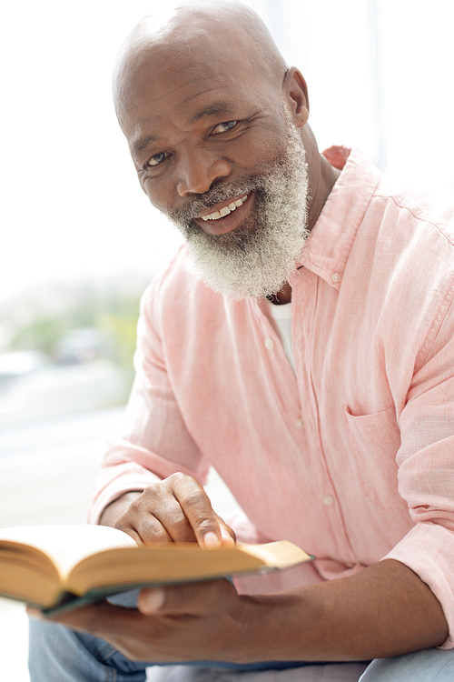 Side view of African American man reading a book and sitting on the bed in the bedroom. He is smiling and looking at the camera. Authentic Senior Retired Life Concept