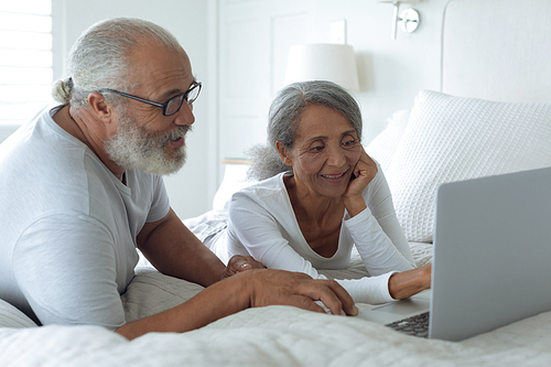 Side view of senior diverse couple sitting on bed inside a room and watching laptop. Authentic Senior Retired Life Concept