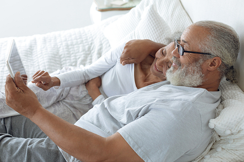 Side view of senior diverse couple lying in bed and watching digital tablet in bedroom. Authentic Senior Retired Life Concept