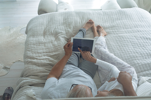 Side view of senior diverse couple lying in bed and watching digital tablet in bedroom. Authentic Senior Retired Life Concept