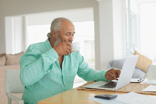 Side view of African-American man using laptop on table and drinking coffee indoor. Authentic Senior Retired Life Concept