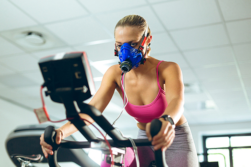 Low angle view of beautiful young Caucasian female athlete with oxygen mask exercising with exercise bike in fitness studio. Bright modern gym with fit healthy people working out and training