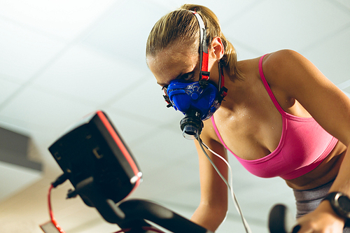 Low angle view of beautiful young Caucasian female athlete with oxygen mask exercising with exercise bike in fitness studio. Bright modern gym with fit healthy people working out and training