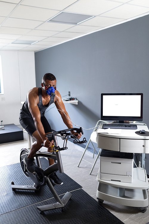 Front view of an African-American athletic man doing a fitness test using a mask connected to a monitor while riding an exercise bike inside a room at a sports center. Athlete testing themselves with cardiovascular fitness test on exercise bike