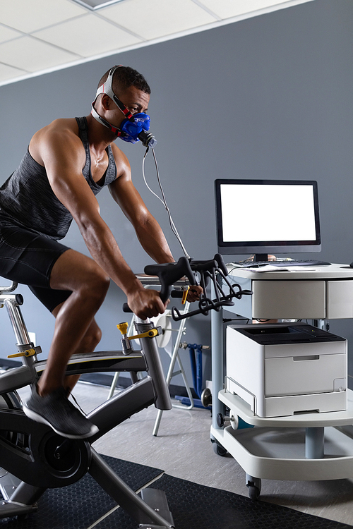 Side view of an African-American athletic man doing a fitness test using a mask connected to a monitor while riding an exercise bike inside a room at a sports center. Athlete testing themselves with cardiovascular fitness test on exercise bike