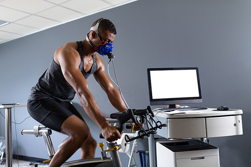 Side view of an African-American athletic man doing a fitness test using a mask connected to a monitor while riding an exercise bike inside a room at a sports center. Athlete testing themselves with cardiovascular fitness test on exercise bike
