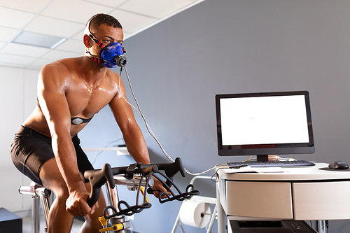 Side view of a naked African-American athletic man doing a fitness test using a mask connected to a monitor while riding an exercise bike inside a room at a sports center. Athlete testing themselves with cardiovascular fitness test on exercise bike