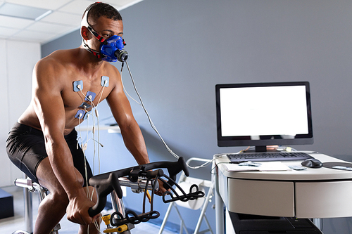 Side view of a naked African-American athletic man doing a fitness test using a mask connected to a monitor while riding an exercise bike inside a room at a sports center. Athlete testing themselves with cardiovascular fitness test on exercise bike
