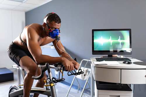 Side view of a naked African-American athletic man doing a fitness test using a mask connected to a monitor with a heart line on the screen while riding an exercise bike inside a room at a sports center. Athlete testing themselves with cardiovascular fitness test on exercise bike