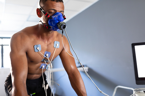 Front view of a naked African-American athletic man doing a fitness test using a mask connected to a monitor while riding an exercise bike inside a room at a sports centre. Athlete testing themselves with cardiovascular fitness test on exercise bike