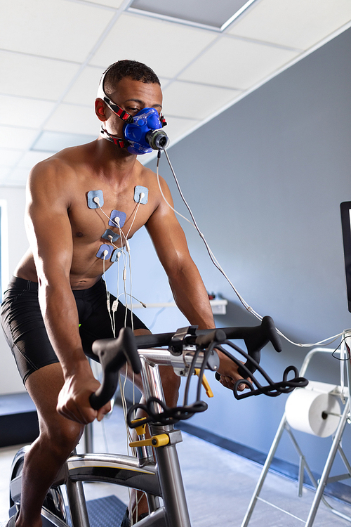 Front view of a naked African-American athletic man doing a fitness test using a mask connected to a monitor while riding an exercise bike inside a room at a sports center. Athlete testing themselves with cardiovascular fitness test on exercise bike