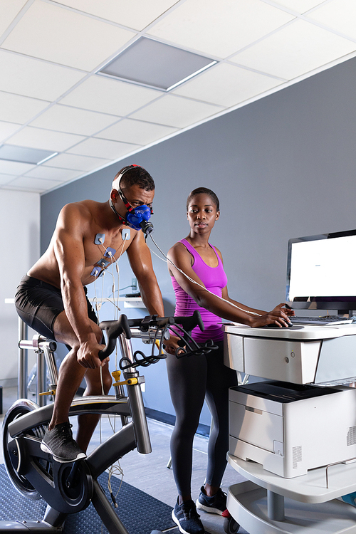 Side view of a naked African-American athletic man doing a fitness test using a mask connected to a monitor while riding an exercise bike and an African-American woman assisting him inside a room at a sports center. Athlete testing themselves with cardiovascular fitness test on exercise bike