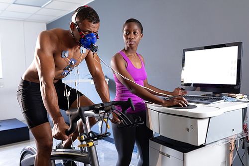 Side view of a naked African-American athletic man doing a fitness test using a mask connected to a monitor while riding an exercise bike and an African-American woman assisting him inside a room at a sports center. Athlete testing themselves with cardiovascular fitness test on exercise bike