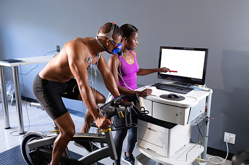 Side view of a naked African-American athletic man doing a fitness test using a mask connected to a monitor while riding an exercise bike and an African-American woman showing him results in the monitor inside a room at a sports center. Athlete testing themselves with cardiovascular fitness test on exercise bike