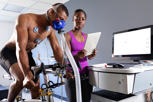 Side view of a naked African-American athletic man doing a fitness test using a mask connected to a monitor while riding an exercise bike and an African-American woman showing him the results inside a room at a sports centre. Athlete testing themselves with cardiovascular fitness test on exercise bike