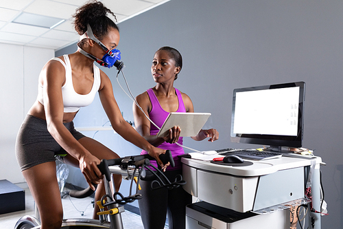 Front view of an African-American athletic woman doing a fitness test using a mask connected to a monitor while riding an exercise bike and an African-American woman monitoring her inside a room at a sports center. Athlete testing themselves with cardiovascular fitness test on exercise bike