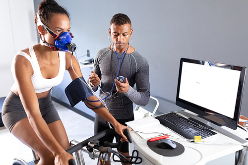 Side view of an African-American athletic woman doing a fitness test using a mask connected to a monitor while riding an exercise bike and an African-American man measuring her blood pressure inside a room at a sports center. Athlete testing themselves with cardiovascular fitness test on exercise bike