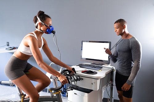Side view of an African-American athletic woman doing a fitness test using a mask connected to a monitor while riding an exercise bike and an African-American man looking at a timer inside a room at a sports center. Athlete testing themselves with cardiovascular fitness test on exercise bike