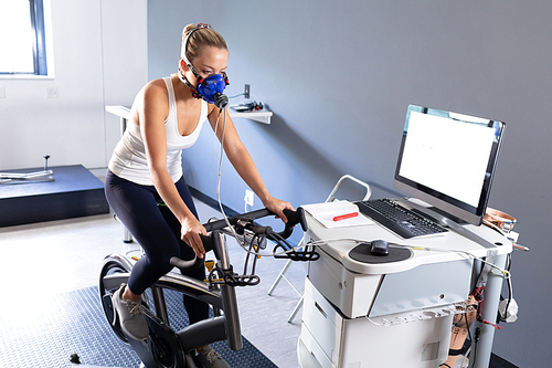 Side view of a Caucasian athletic woman doing a fitness test using a mask connected to a monitor while riding an exercise bike inside a room at a sports center. Athlete testing themselves with cardiovascular fitness test on exercise bike