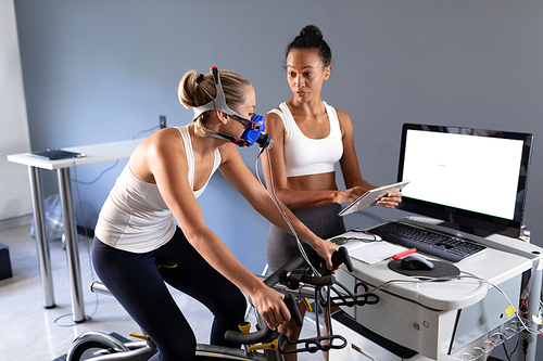 Side view of a Caucasian athletic woman doing a fitness test using a mask connected to a monitor while riding an exercise bike  and an African-American woman monitoring her results inside a room at a sports center. Athlete testing themselves with cardiovascular fitness test on exercise bike