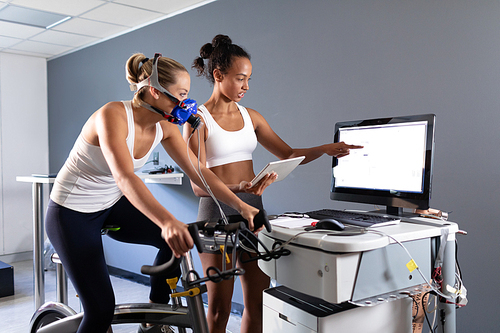 Side view of a Caucasian athletic woman doing a fitness test using a mask connected to a monitor while riding an exercise bike  and an African-American woman explaining her results inside a room at a sports center. Athlete testing themselves with cardiovascular fitness test on exercise bike