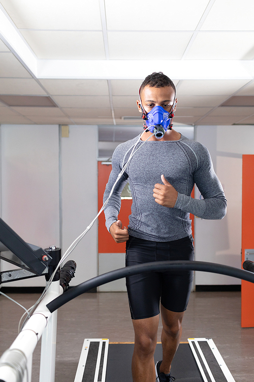 Front view of an African-American athletic man doing a fitness test using a mask connected to a monitor while riding a treadmill inside a room at a sports center. Athlete testing themselves with cardiovascular fitness test on exercise bike