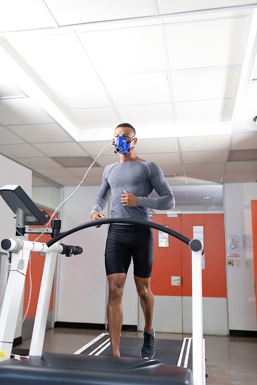 Low angle view of an African-American athletic man doing a fitness test using a mask connected to a monitor while riding a treadmill inside a room at a sports center. Athlete testing themselves with cardiovascular fitness test on exercise bike