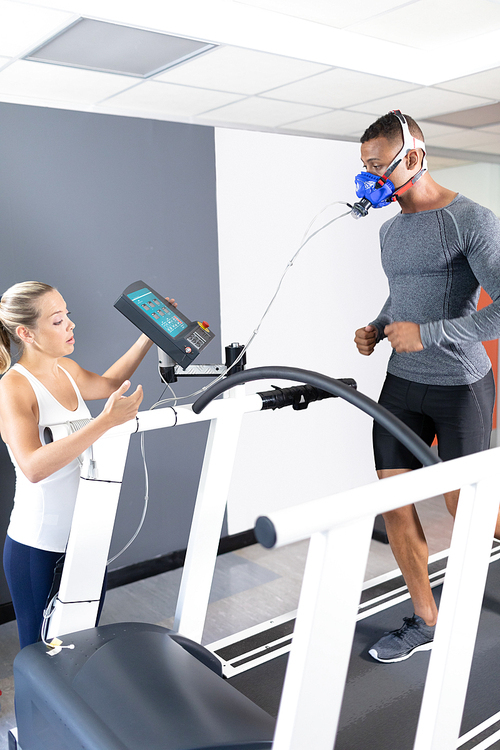 Side view of an African-American athletic man doing a fitness test using a mask connected to a monitor while riding a treadmill and a Caucasian woman assisting inside a room at a sports center. Athlete testing themselves with cardiovascular fitness test on exercise bike