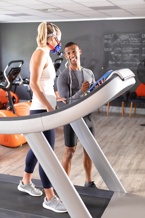 Side view of a Caucasian athletic woman doing a fitness test using a mask  while using a treadmill and an African-American man showing her results inside a room at a sports center. Athlete testing themselves with cardiovascular fitness test on exercise bike