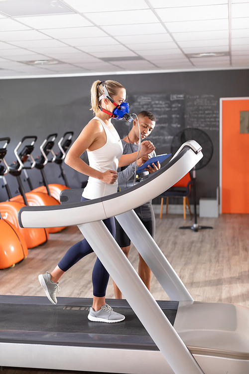 Side view of a Caucasian athletic woman doing a fitness test using a mask  while using a treadmill and an African-American man monitoring her inside a room at a sports center. Athlete testing themselves with cardiovascular fitness test on exercise bike