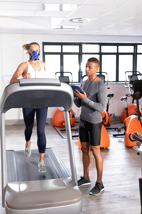 Front view of a Caucasian athletic woman doing a fitness test using a mask while using a treadmill and an African-American man monitoring her inside a room at a sports center. Athlete testing themselves with cardiovascular fitness test on exercise bike