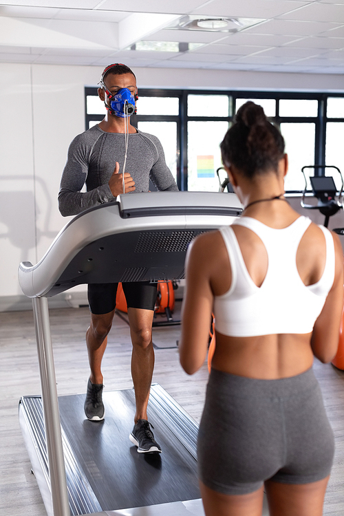 Front view of an African-American athletic man doing a fitness test using a mask while using a treadmill and an African-American woman assisting him inside a room at a sports center. Athlete testing themselves with cardiovascular fitness test on exercise bike