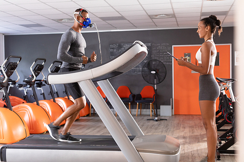 Side view of an African-American athletic man doing a fitness test using a mask while using a treadmill and an African-American woman monitoring him inside a room at a sports center. Athlete testing themselves with cardiovascular fitness test on exercise bike