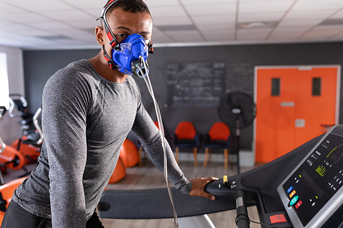 Side view of an African-American athletic man doing a fitness test using a mask while using a treadmill inside a room at a sports center. Athlete testing themselves with cardiovascular fitness test on exercise bike