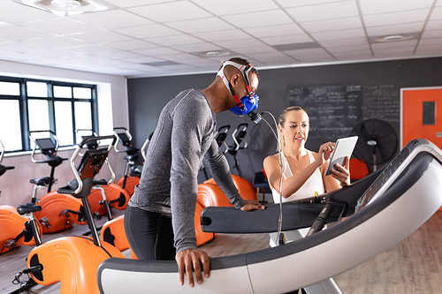 Side view of an African-American athletic man doing a fitness test using a mask while using a treadmill and a Caucasian woman showing his results inside a room at a sports center. Athlete testing themselves with cardiovascular fitness test on exercise bike