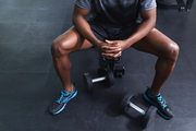 Low section of African-american man sitting with hands clasped on a bench in fitness center. Bright modern gym with fit healthy people working out and training