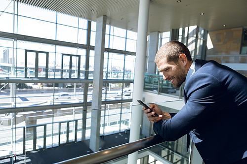 Side view of young Caucasian businessman looking down smiling at his smartphone. He is standing leaning on a handrail in the glass walled atrium of a modern business lobby. Modern corporate start up new business concept with entrepreneur working hard