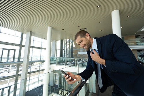 Side view of young Caucasian businessman looking down at his smartphone. He is standing leaning on a handrail in the glass walled atrium of a modern business lobby. Modern corporate start up new business concept with entrepreneur working hard