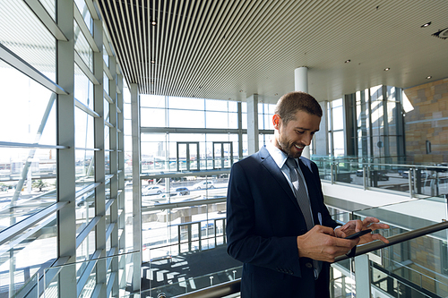 Front view of young Caucasian businessman looking down smiling at his smartphone. He is standing leaning on a handrail in the glass walled atrium of a modern business lobby. Modern corporate start up new business concept with entrepreneur working hard