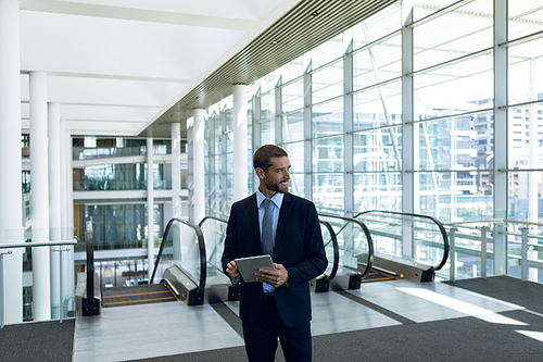 Front view of young Caucasian businessman holding tablet computer and looking away. He is standing in front of escalators in the glass walled lobby of a modern business building. Modern corporate start up new business concept with entrepreneur working hard