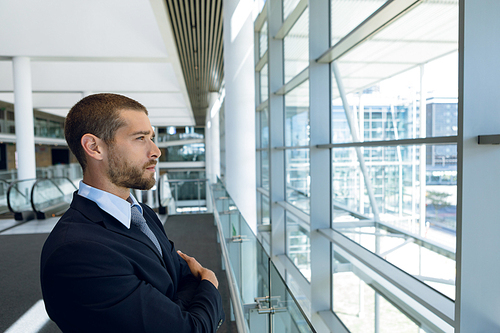 Side view of young Caucasian businessman looking out of window, standing in front of escalotrs in the glass walled lobby of a modern business building. Modern corporate start up new business concept with entrepreneur working hard