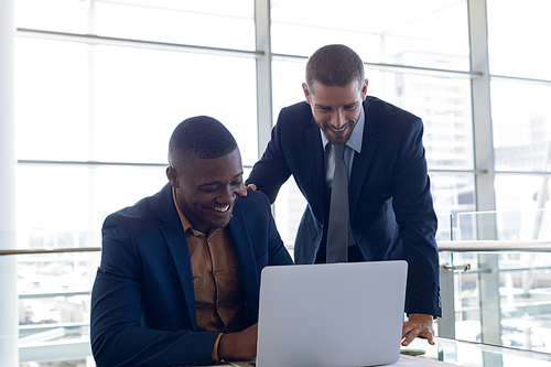 Front view of young African American businessman sitting in front of the window in an office looking at a laptop with a young Caucasian businessman standing beside him with hand on his shoulder. They are both smiling. Modern corporate start up new business concept with entrepreneur working hard