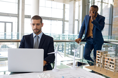 Front view of young Caucasian businessman using laptop sitting at a desk in an architectural office. A young African American businessman is standing using smartphone in the background. Modern corporate start up new business concept with entrepreneur working hard