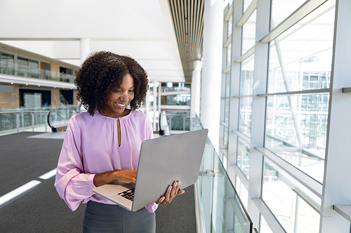 Front view of young African American businesswoman standing using laptop computer in the glass walled lobby of a modern business building. Modern corporate start up new business concept with entrepreneur working hard