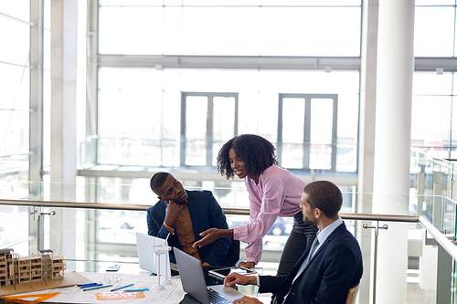 Elevated side view of young African American businesswoman standing at a desk talking with a young Caucasian and an African American businessman sitting using laptops working together in a modern office. Modern corporate start up new business concept with entrepreneur working hard