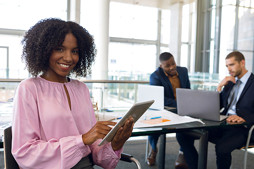 Portrait of young African American businesswoman sitting using tablet computer smiling. Her colleagues working siting at desk in the background. Modern corporate start up new business concept with entrepreneur working hard