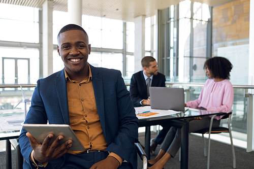 Portrait of young African American businessman sitting holding tablet computer in a modern office smiling to camera. In the background a Caucasian businessman and an African American businesswoman are sitting at a desk using a laptop and talking. Modern corporate start up new business concept with entrepreneur working hard
