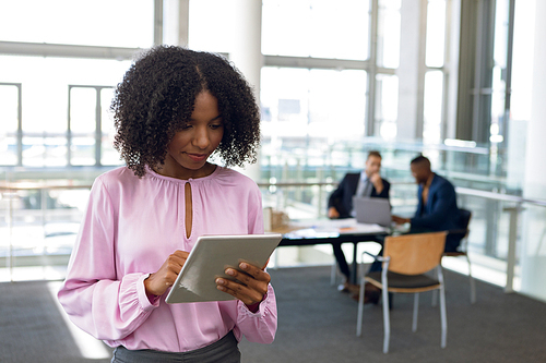 Close up front view of young African American businesswoman standing using tablet computer in a modern office. An African American and a Caucasian businessman work sitting at a desk in the background. Modern corporate start up new business concept with entrepreneur working hard
