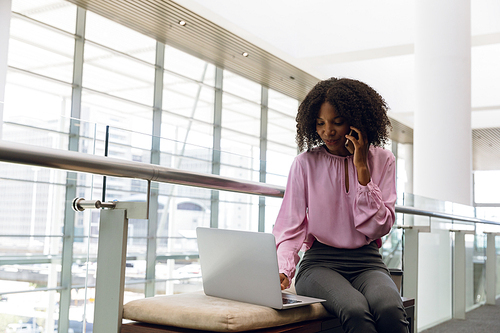 Low angle front view of young African American businesswoman working on a laptop and talking on her phone sitting in the lobby of a modern office. Modern corporate start up new business concept with entrepreneur working hard